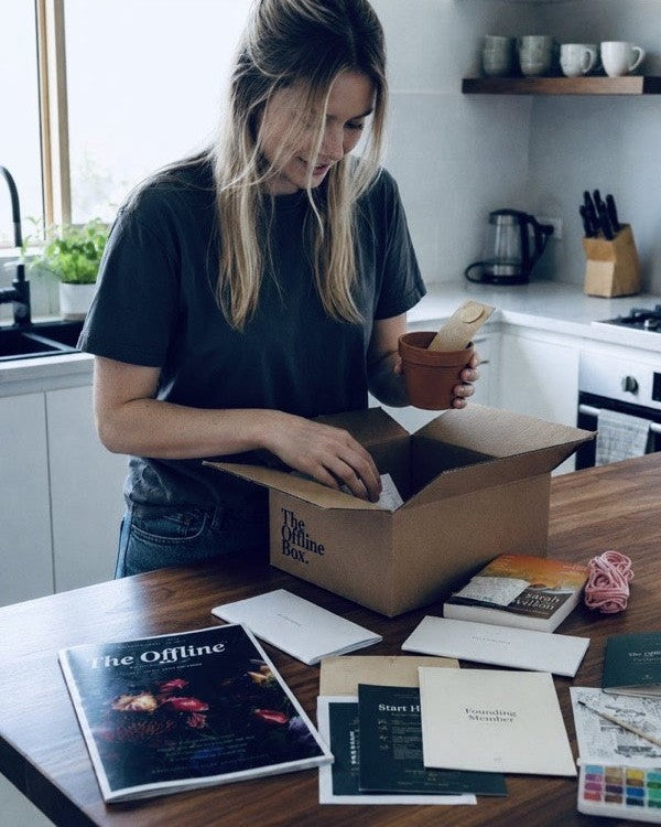 Someone unpacking an Offline Box at a kitchen bench, with the magazine, a book and a terracotta pot on the table